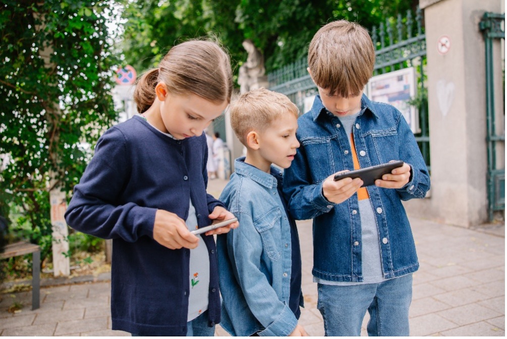 Drei Kinder im öffentlichen Raum blicken auf ihre Smartphones. Ihre Umgebung scheint dabei in den Hintergrund zu rücken.
