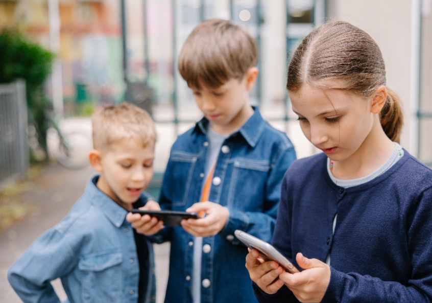 Drei Kinder stehen nebeneinander im öffentlichen Raum und blicken konzentriert auf ihre Smartphones. Ihre Umgebung scheint dabei in den Hintergrund zu rücken.