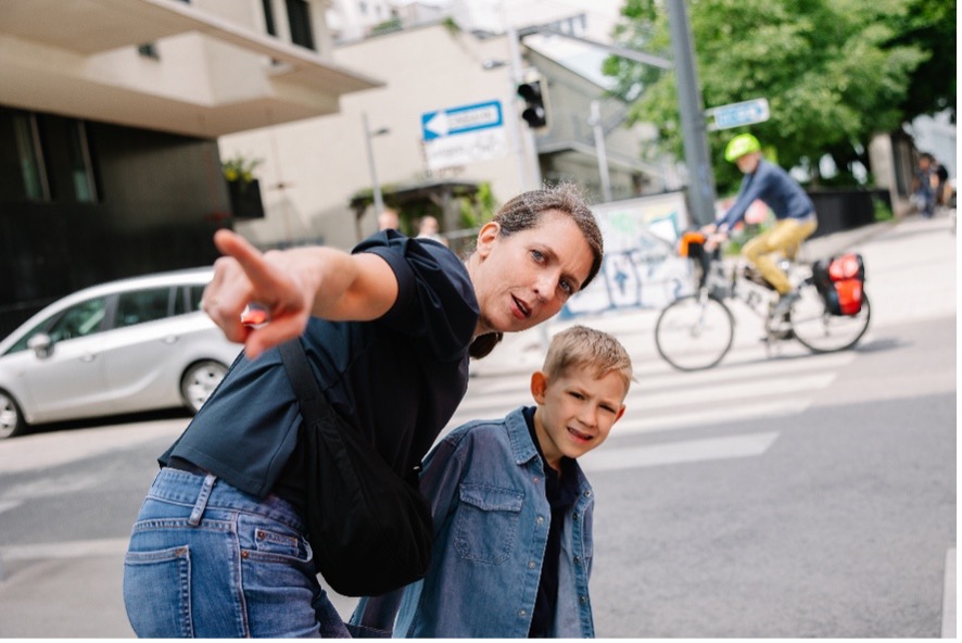 Eine Mutter steht mit ihrem Kind an einer Straßenecke oder am Gehsteig. Sie beugt sich leicht zu ihm herunter und zeigt mit dem Finger in Richtung des Verkehrs. Das Kind schaut aufmerksam dorthin, wo die Mutter hinzeigt.