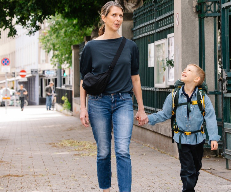Eine Mutter geht mit ihrem Kind an der Hand einen Gehsteig entlang. Das Kind trägt einen Schulranzen – es könnte auf dem Weg zur Schule sein. Die Mutter läuft auf der straßenabgewandten Seite, das Kind auf der sicheren Innenseite des Gehsteigs.