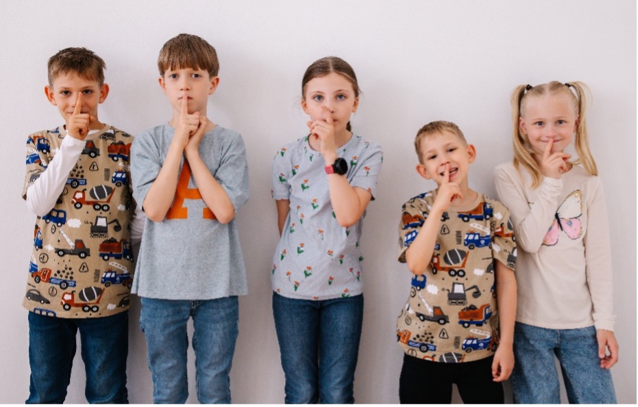 ChatGPT said: Fünf Kinder halten sich den Finger vor den Mund und machen „Pssst“, um aufmerksam den Verkehrsgeräuschen zu lauschen.