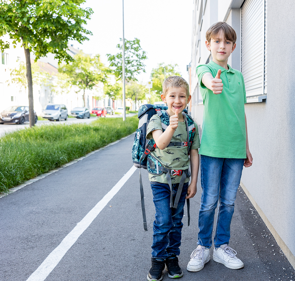 Zwei Kinder unterschiedlichen Alters stehen auf dem Gehsteig und strecken ihre Daumen nach oben.