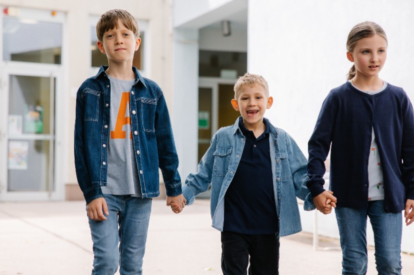 Drei Kinder unterschiedlichen Alters gehen im öffentlichen Raum Hand in Hand; die beiden älteren führen das jüngere Kind.