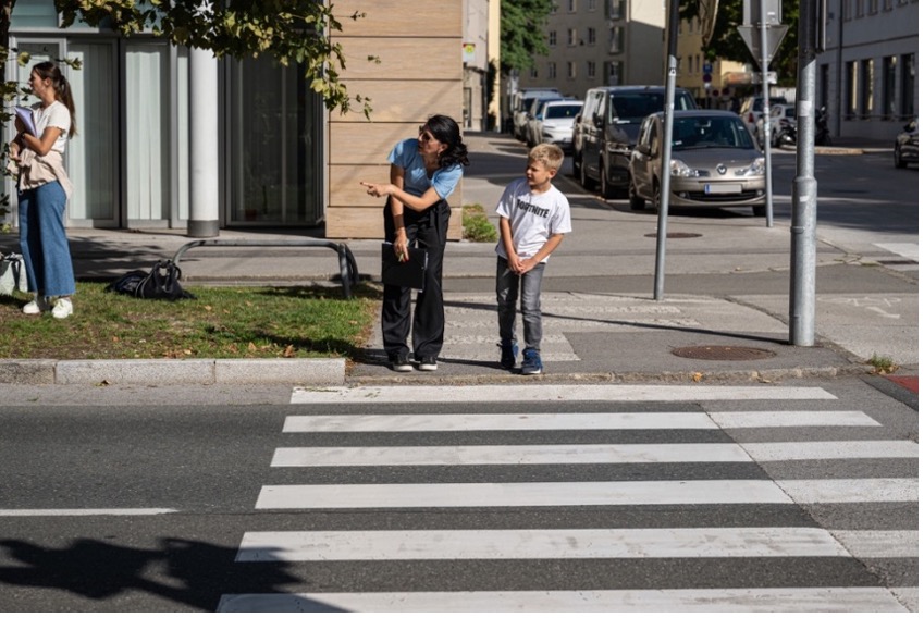 Eine erwachsene Person erklärt einem Kind am Zebrastreifen die richtige Blickrichtung vor dem Überqueren der Straße.