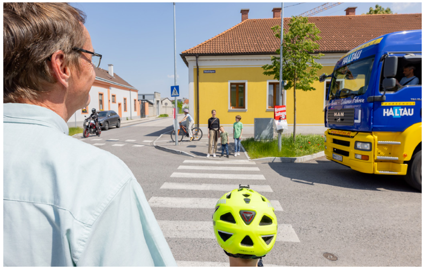 In Abbildung 1 ist folgende Verkehrssituation zu sehen: Ein rollerfahrendes Kind möchte gemeinsam mit einem Erwachsenen eine Fahrbahn überqueren.