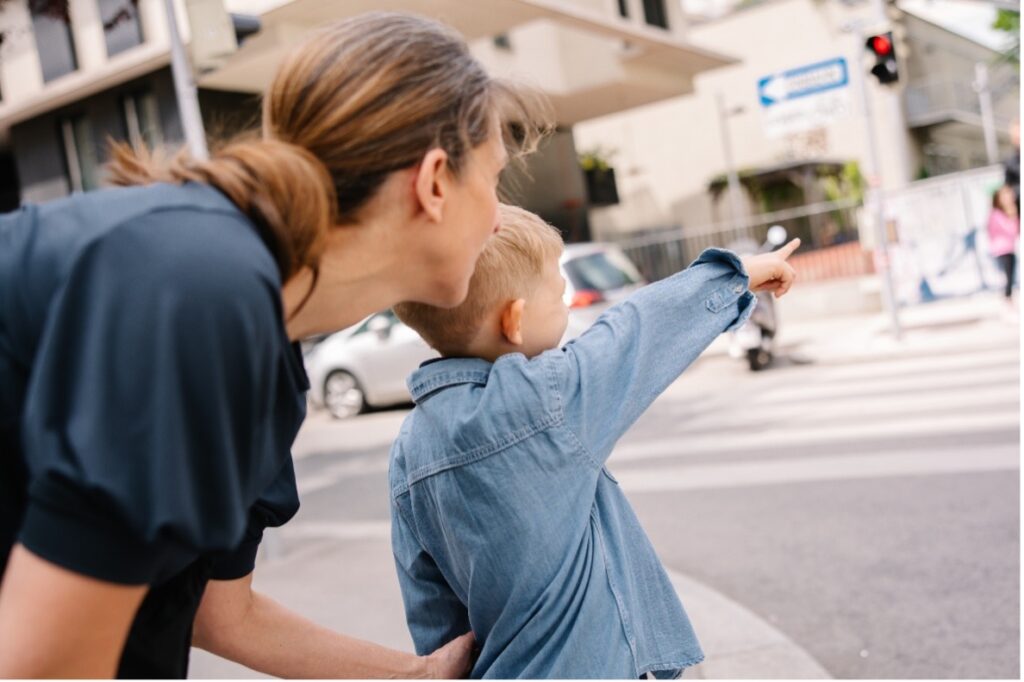 Eine Mutter steht mit ihrem Kind am Zebrastreifen. Das Kind zeigt auf die Autos, während die Mutter das Verkehrsgeschehen erklärt.