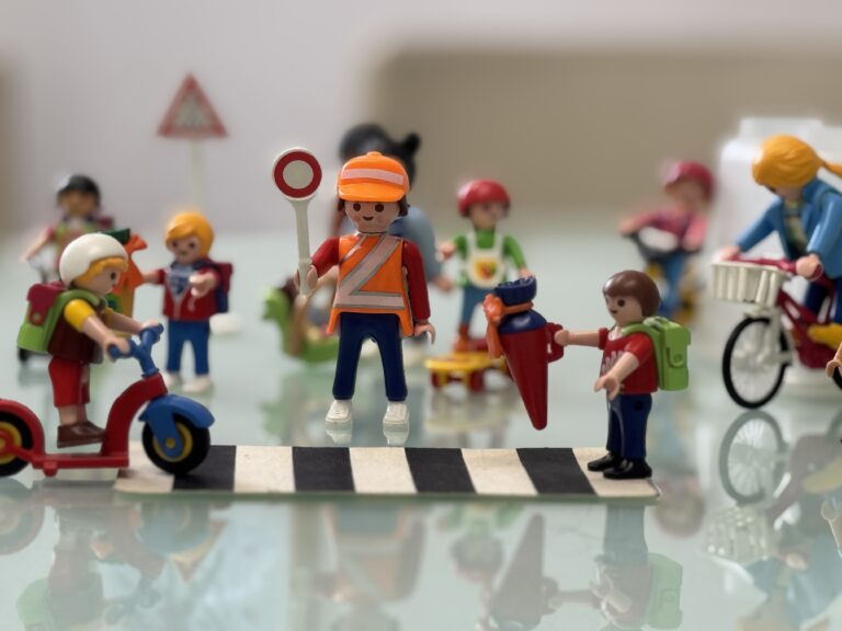 Straßenszene mit Playmobilfiguren: Kinder und verschiedene andere Verkehrsteilnehmer:innen an einem Zebrastreifen