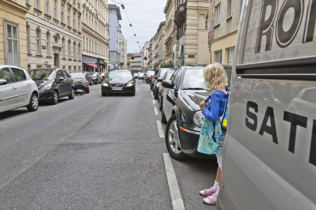 Ein Kind steht zwischen zwei parkenden Autos und schaut auf ein herannahendes Fahrzeug – eine typische, gefährliche Verkehrssituation mit eingeschränkter Sicht.